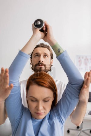 bearded rehabilitation specialist assisting woman exercising with dumbbells in consulting room