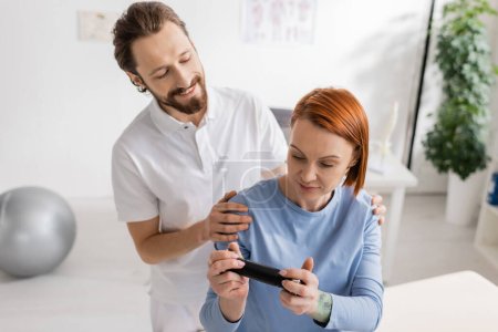 Photo for Positive physiotherapist hugging shoulders of redhead woman looking at mobile phone in consulting room - Royalty Free Image