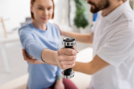 selective focus of dumbbell in hand of woman exercising near blurred physiotherapist in rehabilitation center