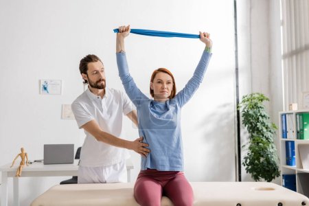 bearded rehabilitation doctor assisting woman sitting on massage table in consulting room and training arms with resistance band