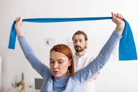 redhead woman working out with elastics near blurred rehabilitologist in recovery center 