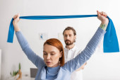 redhead woman working out with elastics near blurred rehabilitologist in recovery center  Sweatshirt #650554908