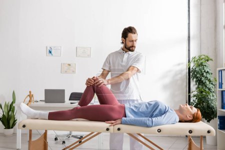 terapeuta manual barbudo haciendo masaje de rodilla a mujer en centro de rehabilitación