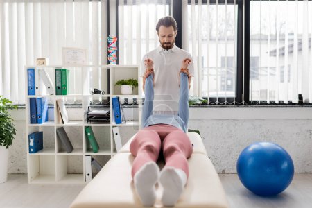 bearded physiotherapist stretching painful arms of woman lying on massage table in modern rehabilitation center