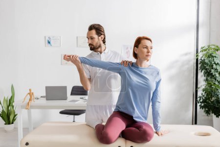 rehabilitologist examining arm of redhead woman sitting on massage table in consulting room