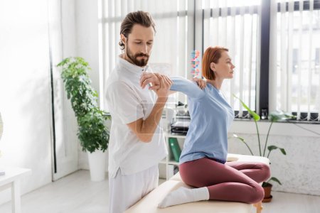 bearded osteopath stretching arm of woman sitting on massage table in rehabilitation center