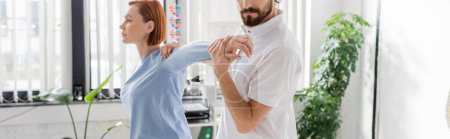 bearded physiotherapist stretching arm of redhead woman while doing diagnostics in consulting room, banner