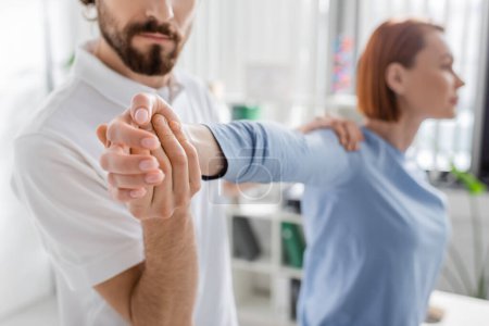 bearded manual therapist examining injured arm of blurred woman in consulting room