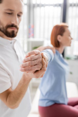 bearded physiotherapist stretching painful arm of blurred woman during diagnostics in consulting room