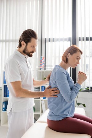 Photo for Side view of bearded physiotherapist touching painful back of redhead woman during appointment in consulting room - Royalty Free Image