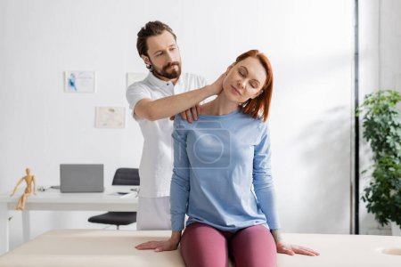 bearded physiotherapist examining painful neck of woman sitting on massage table in consulting room