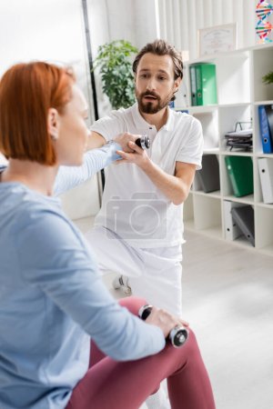 bearded physiotherapist helping redhead woman working out with dumbbells in recovery clinic