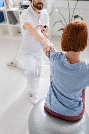 rehabilitólogo brazo de apoyo de mujer pelirroja sentada en la pelota de fitness y haciendo ejercicio con la mancuerna en la sala de consulta