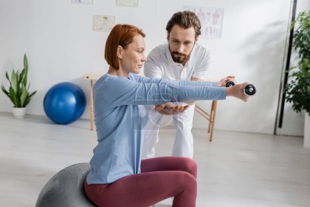 bearded physiotherapist assisting redhead woman sitting on fitball and training with dumbbells in rehabilitation center