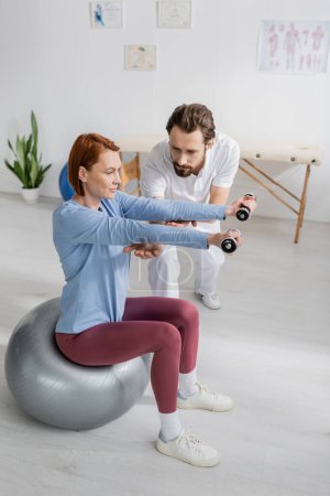 rehabilitologist helping redhead woman working out with dumbbells while sitting on fitness ball in rehab center