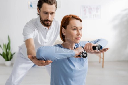 rehabilitologist assisting redhead woman working out with dumbbells in recovery center