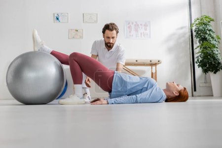 Frau liegt am Boden und trainiert mit Fitball in der Nähe des bärtigen Physiotherapeuten im Reha-Zentrum