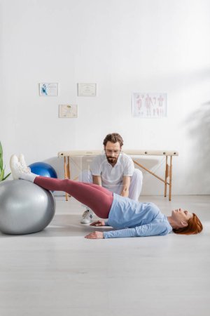 bärtige Physiotherapeutin neben Frau, die am Boden liegt und im Reha-Zentrum mit Fitnessball trainiert