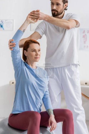 physiotherapist supporting arm of redhead woman training on fitness ball in recovery clinic