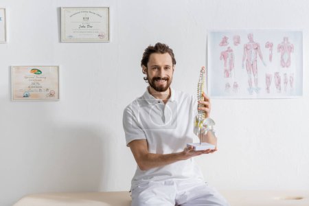 joyful chiropractor smiling at camera while sitting with spine model in rehab center