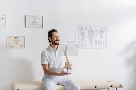 Foto de Fisioterapeuta alegre con modelo de columna vertebral mirando hacia otro lado mientras está sentado en la mesa de masaje en el centro de rehabilitación - Imagen libre de derechos