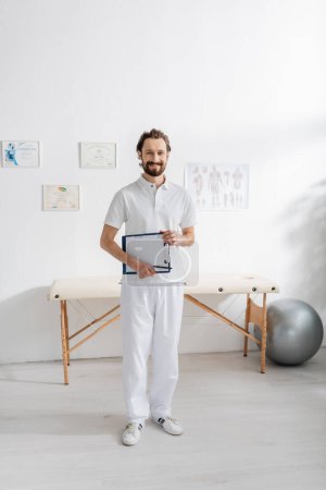 Photo for Full length of carefree physiotherapist holding clipboard with paper while standing in consulting room - Royalty Free Image