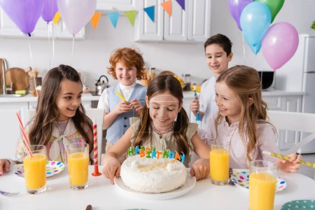 Foto de Chica feliz mirando su pastel de cumpleaños con velas cerca de amigos durante la celebración en casa - Imagen libre de derechos