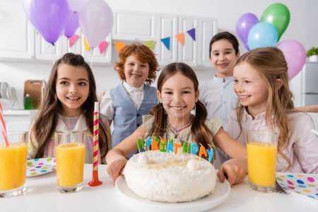 niños felices mirando a la cámara cerca de pastel de cumpleaños y globos durante la celebración en casa 