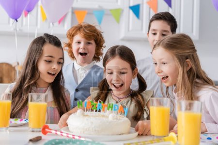 Gruppe verblüffter Kinder betrachtet Geburtstagstorte mit Kerzen bei Heimparty 