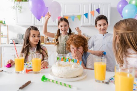 Foto de Pelirroja cubriendo la cara mientras mira pastel de cumpleaños cerca de amigos durante la fiesta en casa - Imagen libre de derechos
