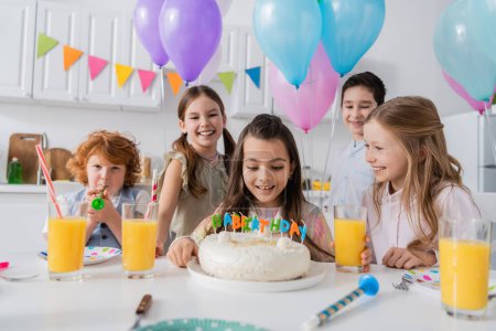 fröhliches Mädchen blickt auf Geburtstagstorte in der Nähe einer glücklichen Gruppe von Freunden während einer Party zu Hause  