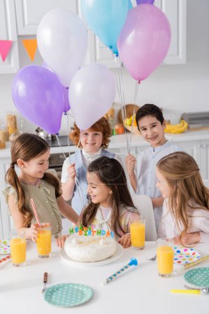 Foto de Niños alegres con globos mirando chica de cumpleaños cerca de la torta y cuernos de fiesta en la mesa - Imagen libre de derechos