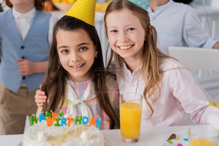 happy girl hugging cheerful friend in party cap next to birthday cake 