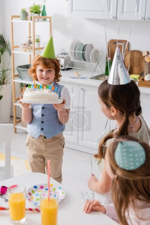 Téléchargez les photos : Heureux rousse garçon tenant gâteau d'anniversaire avec des bougies près des amis pendant la fête à la maison - en image libre de droit