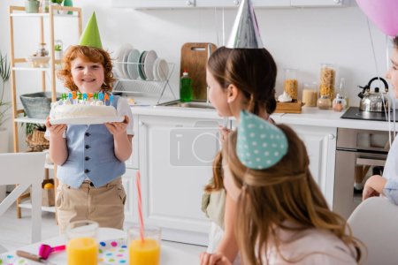 Téléchargez les photos : Heureux rousse garçon tenant gâteau d'anniversaire avec des bougies près des amis joyeux pendant la fête à la maison - en image libre de droit