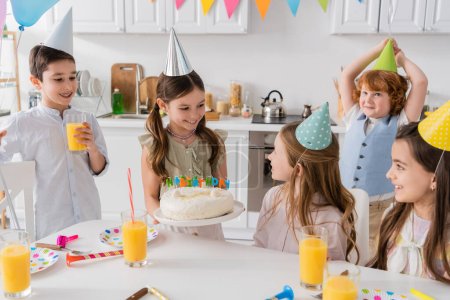 Téléchargez les photos : Fille heureuse tenant gâteau d'anniversaire avec des bougies près des amis gais pendant la fête à la maison - en image libre de droit