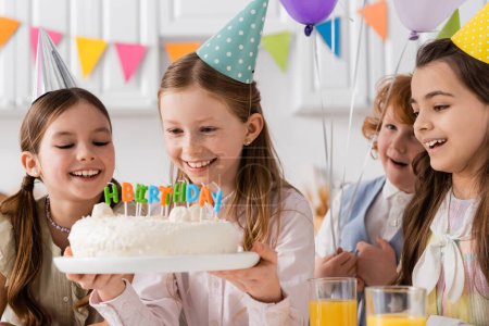 chica feliz celebración de pastel de cumpleaños con velas cerca de niñas alegres durante la celebración en casa 