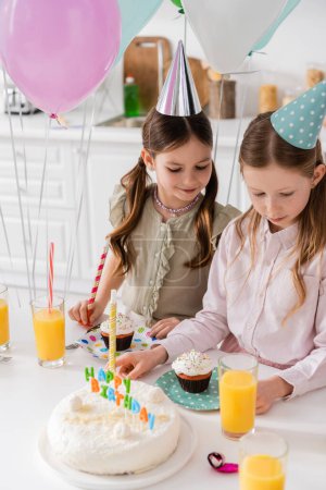 preteen girls in party caps looking at birthday cake and cupcakes on table 