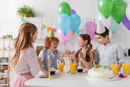 Téléchargez les photos : Filles heureuses dans les casquettes de fête applaudissant les mains et chantant chanson joyeux anniversaire près des garçons et gâteau - en image libre de droit