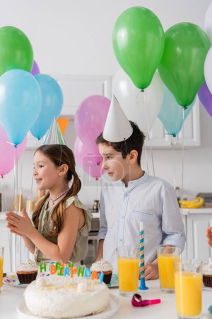 Foto de Alegre chica y chico de pie cerca de pastel de cumpleaños con velas al lado de globos - Imagen libre de derechos