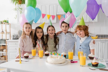 Positive Kindergruppe singt Happy Birthday Song neben Kuchen mit Kerzen und Luftballons 