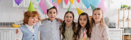 positive group of children singing happy birthday song next to cake with candles and balloons, banner 