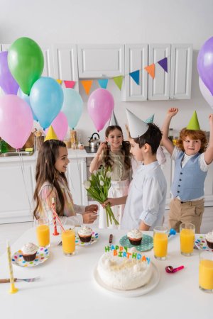 preteen boy giving flowers to cheerful birthday girl near friends on blurred background 