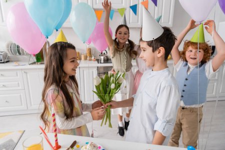 Photo for Happy boy giving tulips to cheerful birthday girl near friends on blurred background - Royalty Free Image