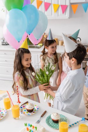 preadolescente chico dando tulipanes a alegre cumpleaños chica cerca de niños en fondo borroso 