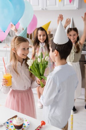 Foto de Feliz chica con vaso de jugo tomando tulipanes de chico cerca de amigos en fondo borroso - Imagen libre de derechos