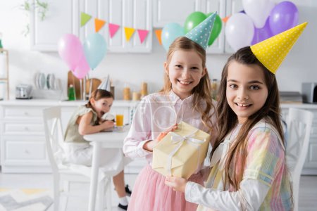 Photo for Cheerful birthday girl in party cap receiving present from friend while looking together in camera - Royalty Free Image
