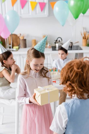 amazed girl in party cap receiving birthday present from redhead boy 
