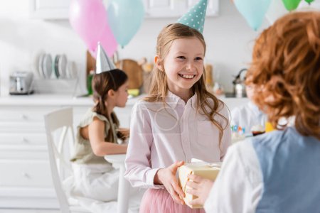 glückliches Mädchen mit Mütze erhält Geschenk von Rotschopf-Freund während Geburtstagsfeier 
