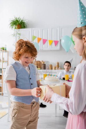 preteen girl in party cap giving present to cheerful redhead boy during birthday celebration 